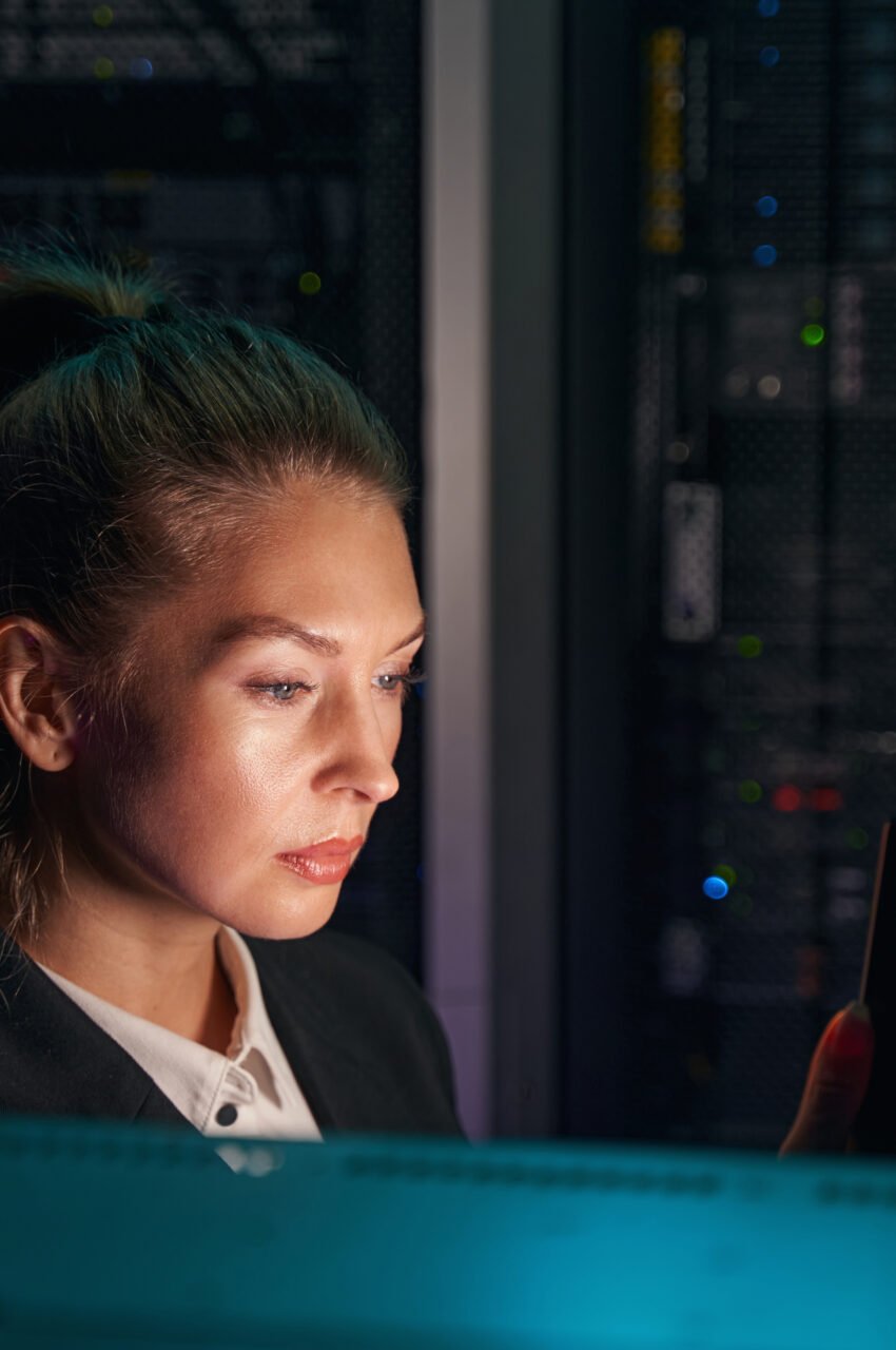 Photo of female network technician configuring servers by using cellphone in dark room of data center